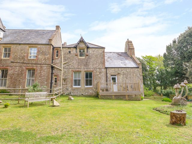 An exterior view of a stone house with a wooden deck and bench in a grassy yard with trees at The Old Kitchens in Totland Bay