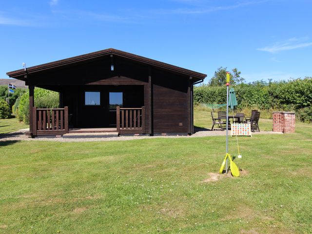 A wooden cabin with a covered porch on a grassy lawn with outdoor furniture and a backyard game at Maple Lodge Lanivet near Bodmin