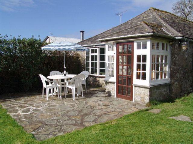 A stone patio with a white table and chairs and a striped umbrella outside a cottage with multiple windows and a red door at Tumrose Cottage in Blisland