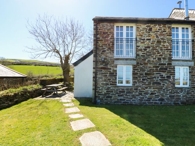 A stone house with white windows and a small patio with a wooden picnic table and bench in a grassy garden at Atlantic View in Tintagel