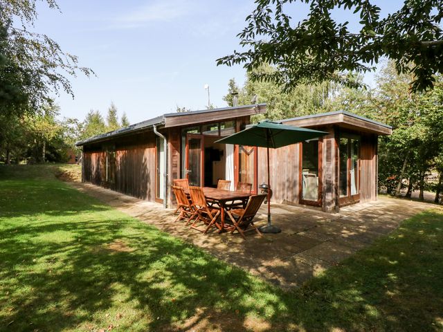 A wooden house with patio doors and outdoor dining set with umbrella on a paved area surrounded by grass and trees at Seaton in Exton Rutland