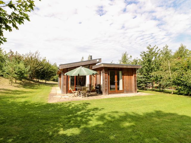 A small wooden house with a patio table and umbrella on a grassy lawn surrounded by trees at Tickencote in Exton Rutland