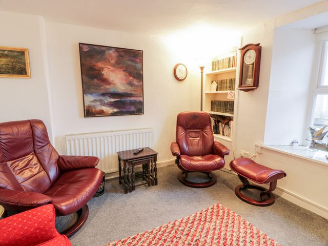 A sitting area with two red leather chairs a footstool small wooden tables a bookshelf wall clock and a window at Sea Spray in Eyemouth