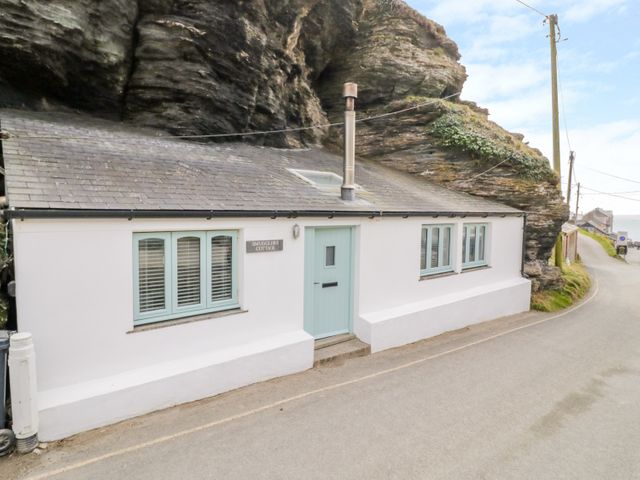 A white cottage with a blue door and windows built into a rocky cliff beside a road at Smugglers Cottage Trebarwith Strand near Tintagel