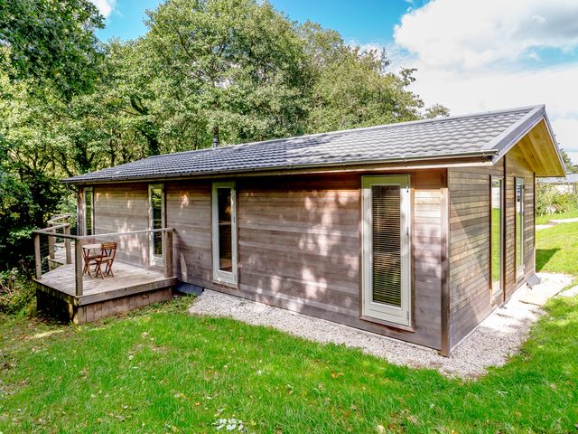 A wooden cabin with small deck and two doors surrounded by grass and trees at 8 Streamside in Lanreath