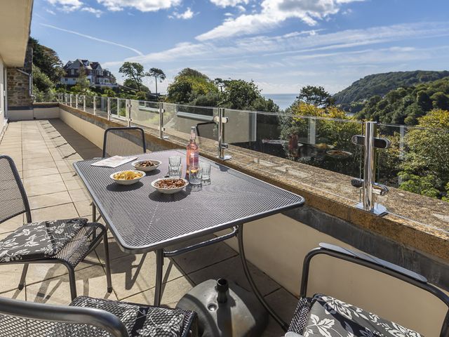 A balcony with a metal table and chairs with cushions and snacks with a glass railing overlooking trees and hills at 5 Fairhaven in Salcombe