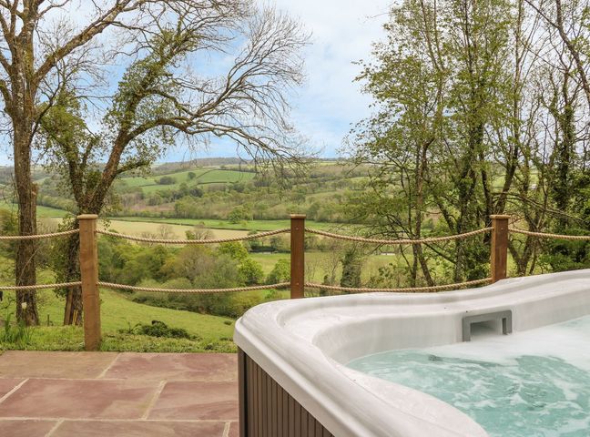 An outdoor hot tub on a stone patio overlooking a fenced green countryside at Little Silver Leaf in High Bickington