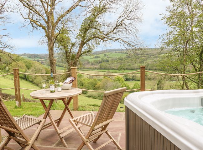 An outdoor patio with two wooden chairs and a table with a bottle of wine and glass next to a hot tub overlooking green hills at Little Silver Owl in High Bickington