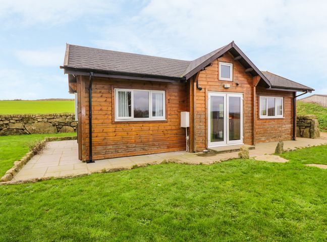 A wooden cabin with white framed windows and doors on a stone patio surrounded by grass at Borah Lodge in Mousehole