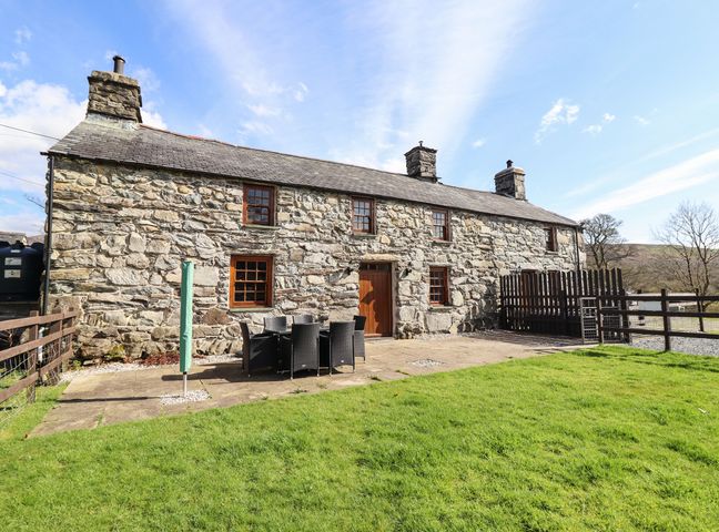 An outdoor area with a stone cottage having multiple windows and chimneys with a patio table and chairs on a paved area at Cwm Yr Afon Mawr in Llanbedr near Harlech