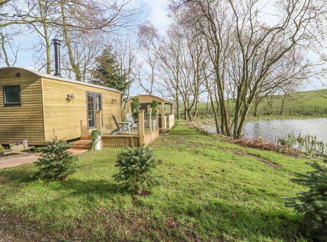A wooden cabin with a small deck and chairs near trees and a body of water at High Rigg Shepherd's Delight in Brampton Cumbria