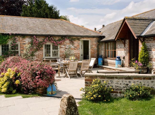 A brick and flint house with a slate roof and an outdoor patio area with wooden table and chairs surrounded by flowering plants at Churn House in Dewlish