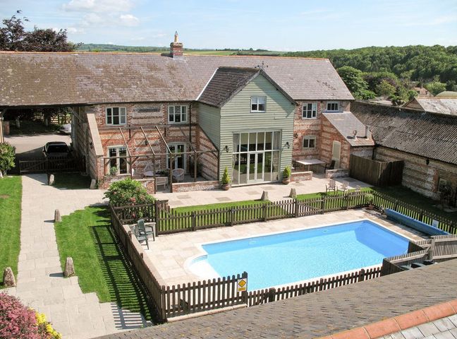 A fenced swimming pool with chairs and lawn in front of a brick and wood house at Manor Farm Barn in Dewlish