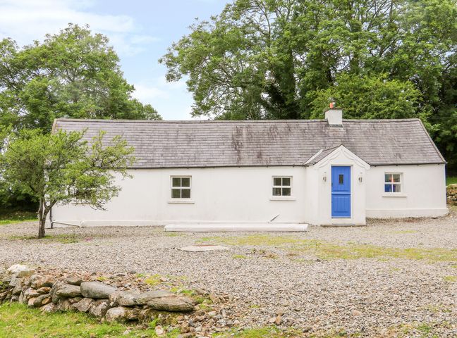 A white cottage with a blue door and three windows surrounded by trees and a gravel yard at The Old White Cottage in Shillelagh County Wicklow