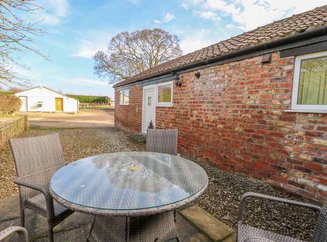 A patio area with a round glass table and four grey woven chairs next to a brick building at The Cottage at Manor Farm in York