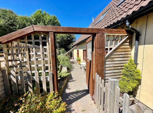 A wooden gate opening to a paved walkway beside a beige building with plants and wooden fencing surrounding the area at Mendip Cottage in Shipham