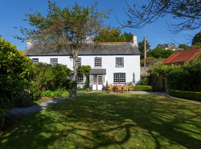 A white two-story house with black window frames and a garden with a tree and outdoor wooden furniture at Court Prior Cottage in Cornworthy