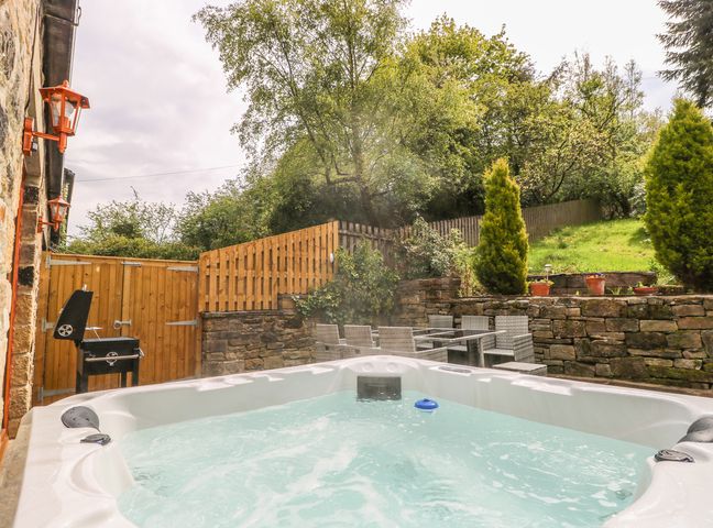 An outdoor area with a hot tub a black barbecue grill a glass table with chairs and a stone wall at Middle Barn in Cross Roads near Haworth