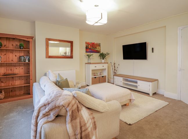 A living room with a sofa an ottoman a wooden bookshelf a wall-mounted tv and a cabinet at Beside The Sea - In Town in Llandudno
