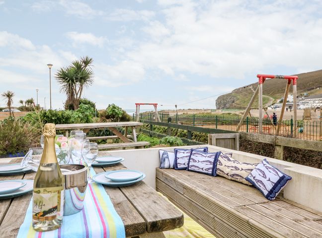 An outdoor dining area with a wooden table set with plates glasses a bottle and cushioned bench seating near a beach with swings at Offshore in Porthtowan
