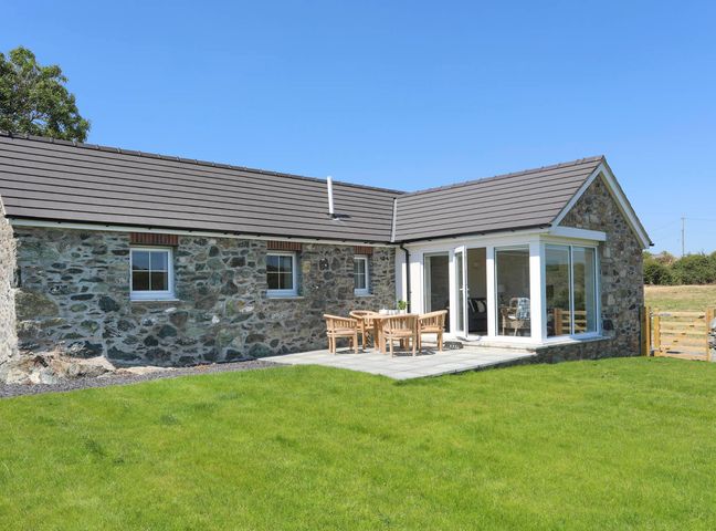 A stone house with three windows and a patio with wooden chairs and a table on a lawn at Bwthyn Glanrhyd in Llanfair yn Neubwll