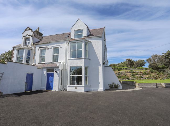 A white two-story house with large windows and a blue door with a paved driveway and garden area at Bryn Dale in Trearddur Bay