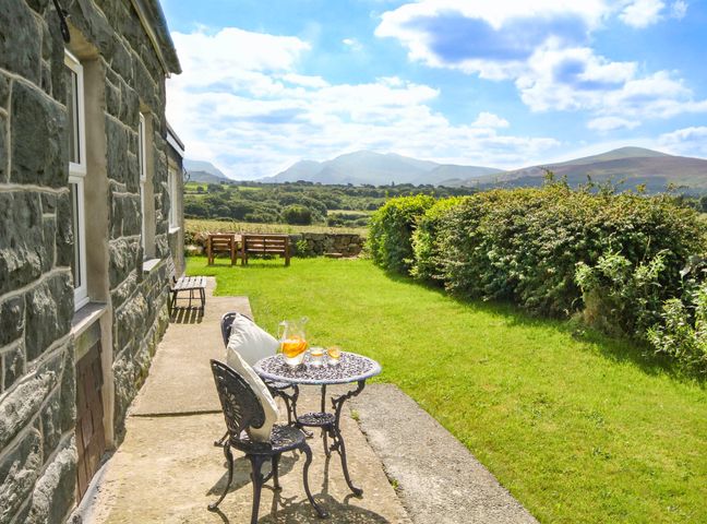 A stone house with a small metal table and two chairs on a patio overlooking grass and hills at Coed Bolyn Lodge in Bethel