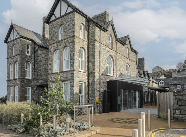 A stone building with arched windows and a modern entrance with glass doors at Harlech Apartments Matholwch in Harlech