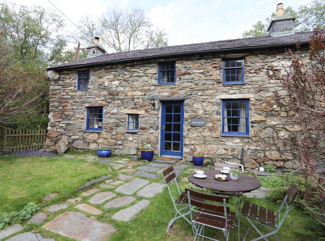 A stone cottage with blue window frames and door a stone pathway and outdoor table with chairs in a garden at Llwyn Bedw in Nant Peris