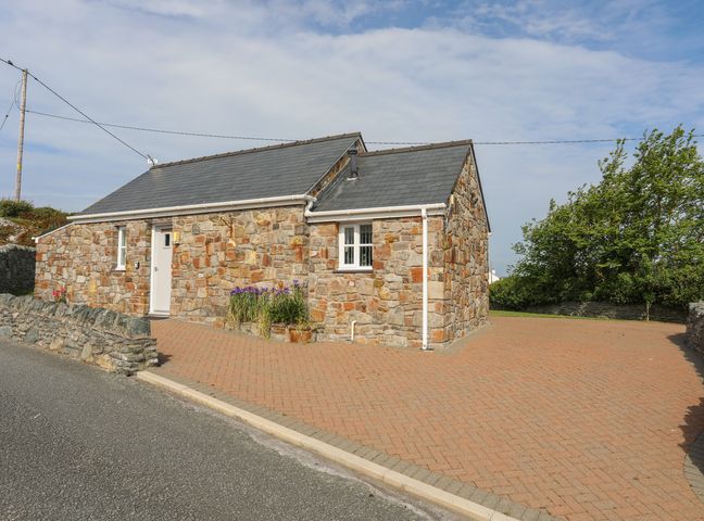 A stone house with a slate roof a white door and windows a brick driveway and some plants at Tyn Towyn - Bwthyn Carreg in Trearddur Bay