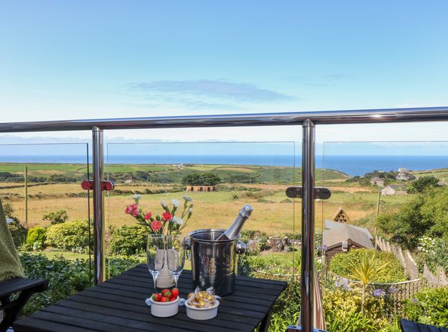 A balcony with a table holding flowers glasses a bucket with a bottle and bowls of strawberries and chocolates overlooking fields and the sea at Sunset in St Just