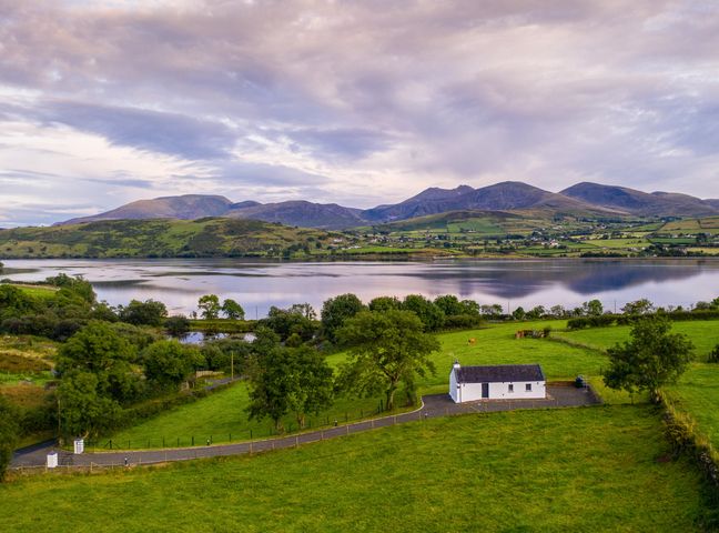 A rural landscape with a small white house near a lake and green fields with mountains in the background at Lough Island Reavy Cottage Kilcoo Mourne Mountains Newry County Down