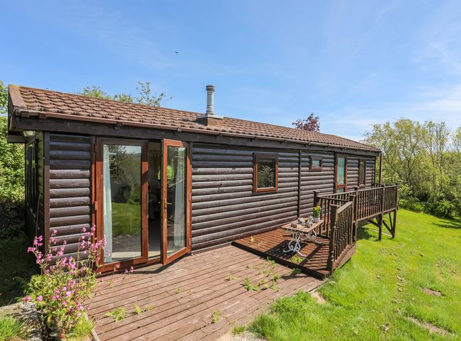 A wooden chalet with a small deck and outdoor table set in a grassy area at Castell Chalet in Penrhos near Pwllheli