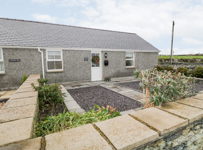 A stone wall and garden with shrubs in front of a grey pebble dash house with a tiled roof at Ysgubor Llwyd in Valley