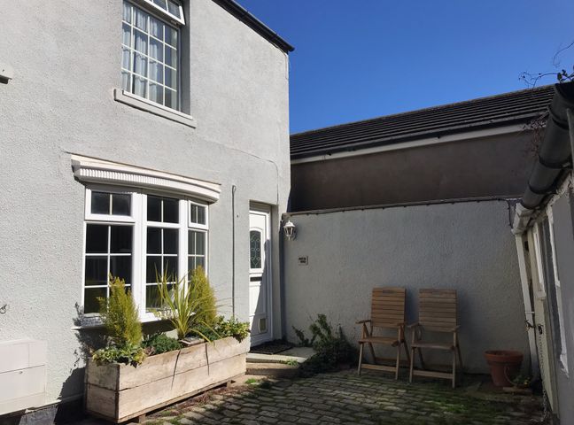 An outdoor courtyard with two wooden chairs a flower planter under a window and a white door at Witts End in Marske-By-The-Sea
