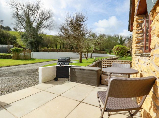 A patio with a table and two chairs a barbecue grill and storage box outside a stone building at Rose Bud Cottage in Polgooth