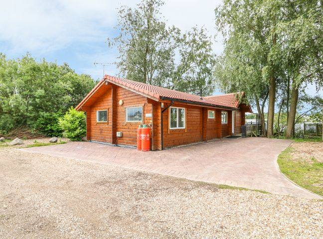 A wooden cabin with a tiled roof surrounded by trees and a gravel driveway at Teal Lodge in Tattershall
