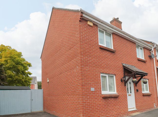 A red brick semi detached house with white windows and a white door with a wooden canopy and hanging flower baskets at Pebbles Foxglove Way in West Bay
