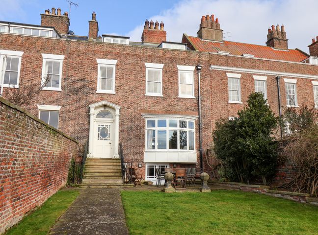 A brick terraced house with white framed windows a pathway leading to a door and a garden with outdoor furniture at 12 St Hildas in Whitby