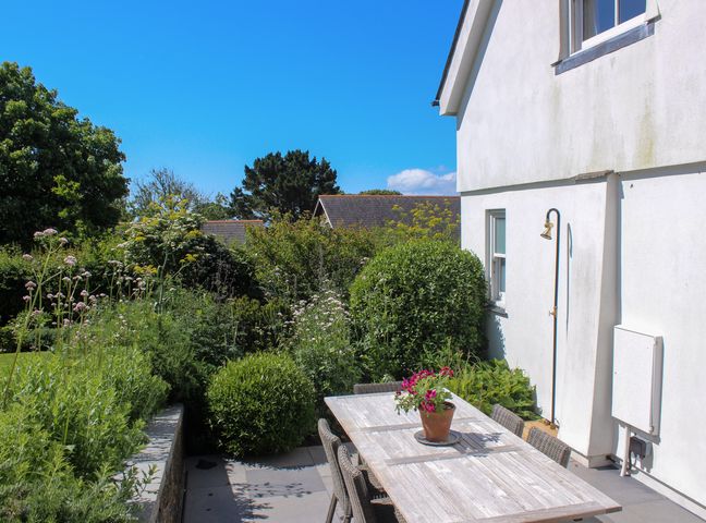 A patio with a wooden dining table and chairs next to a white house with a garden surrounding it at Willow Cottage in Dartmouth