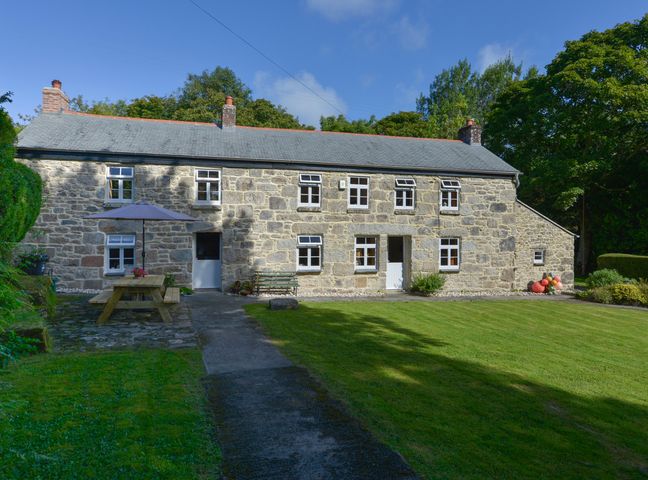 A stone cottage with white framed windows and doors a picnic table with an umbrella and green lawn at Heather Cottage in Nancledra