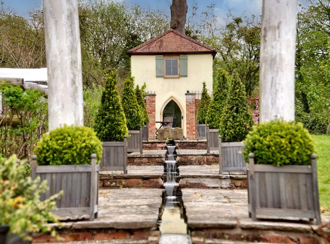 A garden with a small building at the end of a stone pathway with water features and potted plants at The Folly in Weobley