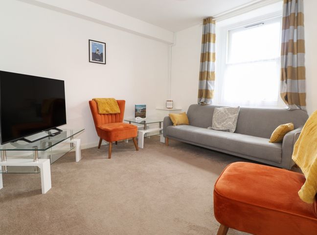 A living room with a large window with striped curtains a gray sofa with cushions two orange chairs a television on a glass stand and a small glass side table at Holly Cottage in Keswick