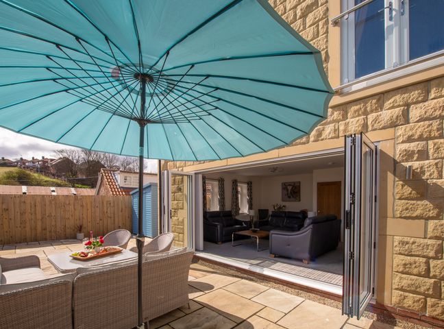 An outdoor patio with a large blue umbrella a wicker seating set and a table with food next to a living room with black leather sofas at Lemonade Cottage in Whitby