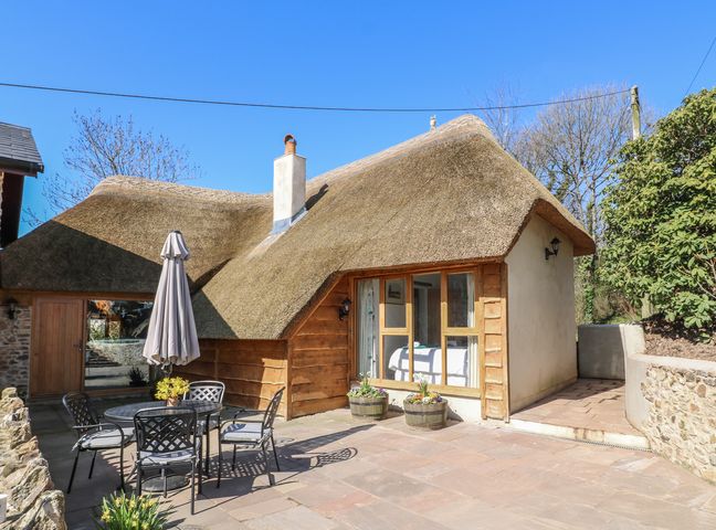 An outdoor patio area with a table umbrella and chairs next to a thatched roof cottage at The Old Smithy in Luppitt