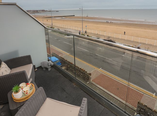 A balcony with two wicker chairs and a table overlooking a road and a sandy beach at Puffin View in Rhos-On-Sea