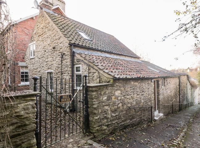 A stone house with a tiled roof behind a black metal gate labeled Hill House on a sloped stone pathway at Dovecote in Pickering