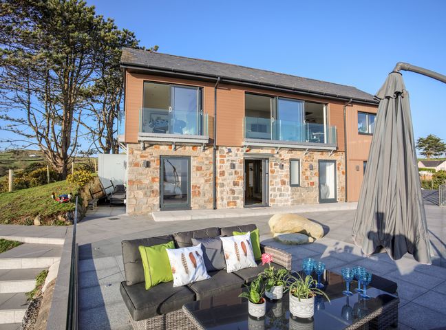 An outdoor patio with a sofa, table with plants and glasses, umbrella, and a two-story house with stone and wood exterior at Hafod Y Mor in Criccieth