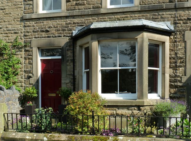 A stone house exterior with a red door and a bay window with plants and flowers in front at Church View in Buxton