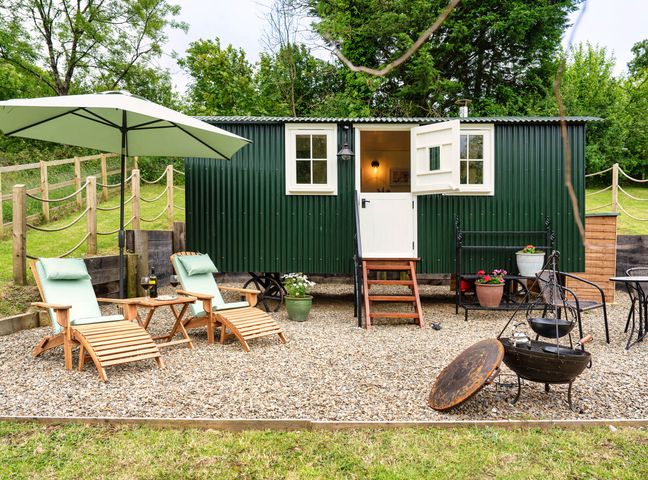 A green metal shepherd hut with white windows and door on a gravel patio with two wooden lounge chairs under a large umbrella and a fire pit in front at Onnen Oakford near Aberaeron
